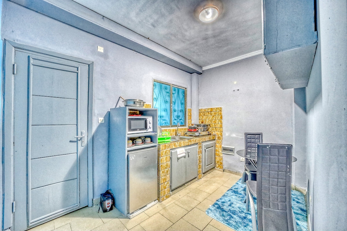 A compact kitchen area features modern appliances, including a refrigerator and kitchen cabinets. A textured blue rug covers the floor, while yellow-tiled backsplash adds color to the countertop. A window allows natural light to brighten the space.