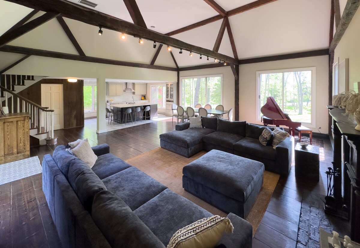 A spacious living area is highlighted by high, wooden-beamed ceilings and large windows, allowing ample natural light. Comfortable seating is arranged around a central area rug, complemented by a grand piano in one corner and a dining area visible in the background.
