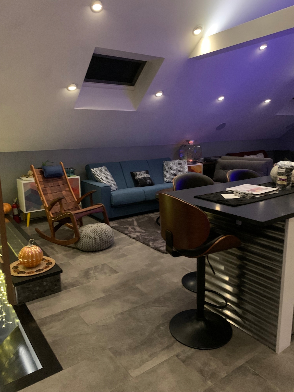 An inviting loft area is showcased with modern decor and ambient lighting. A blue sofa is paired with decorative pillows, while a sleek bar counter features two high stools. A cozy chair and a decorative pouf add comfort to the space, complemented by soft lighting and a large skylight.