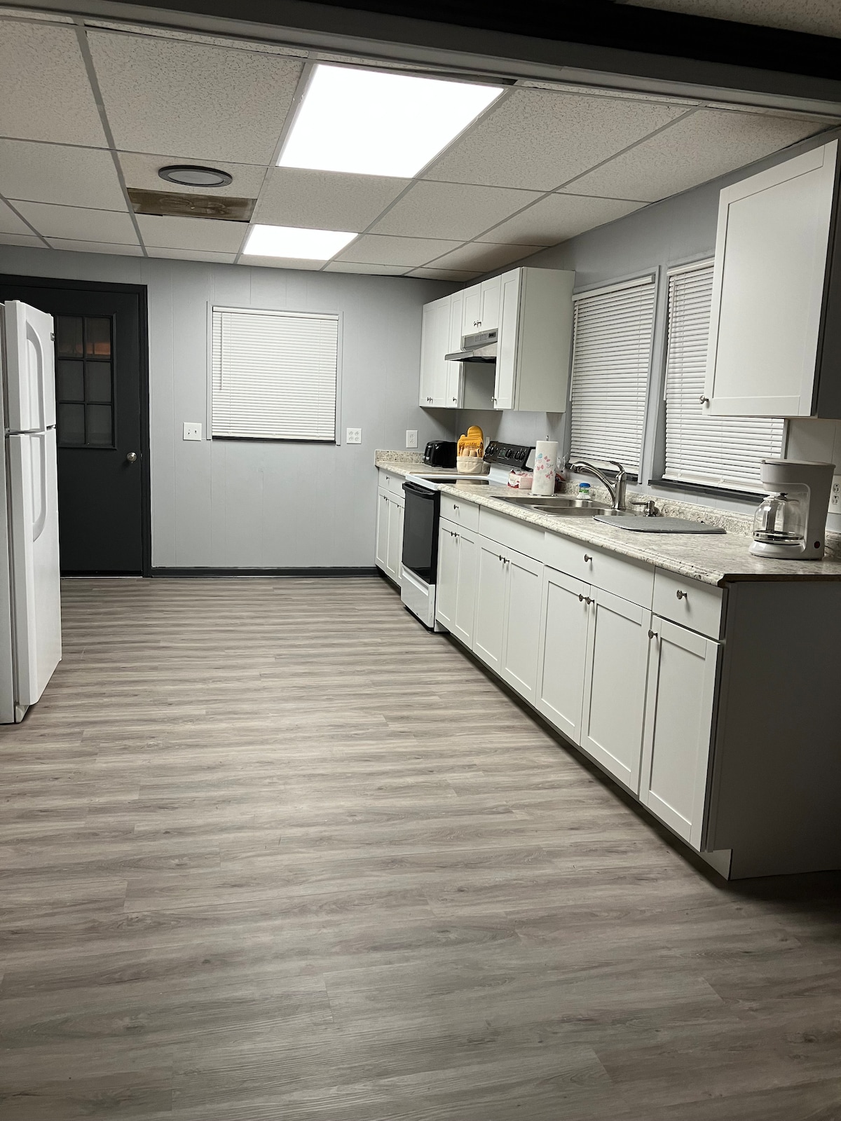The kitchen features modern white cabinetry and a light countertop, complemented by stainless steel appliances. Ample natural light enters through the window, enhancing the clean and spacious layout. A refrigerator and stove are visible, along with a sink positioned beneath the window.