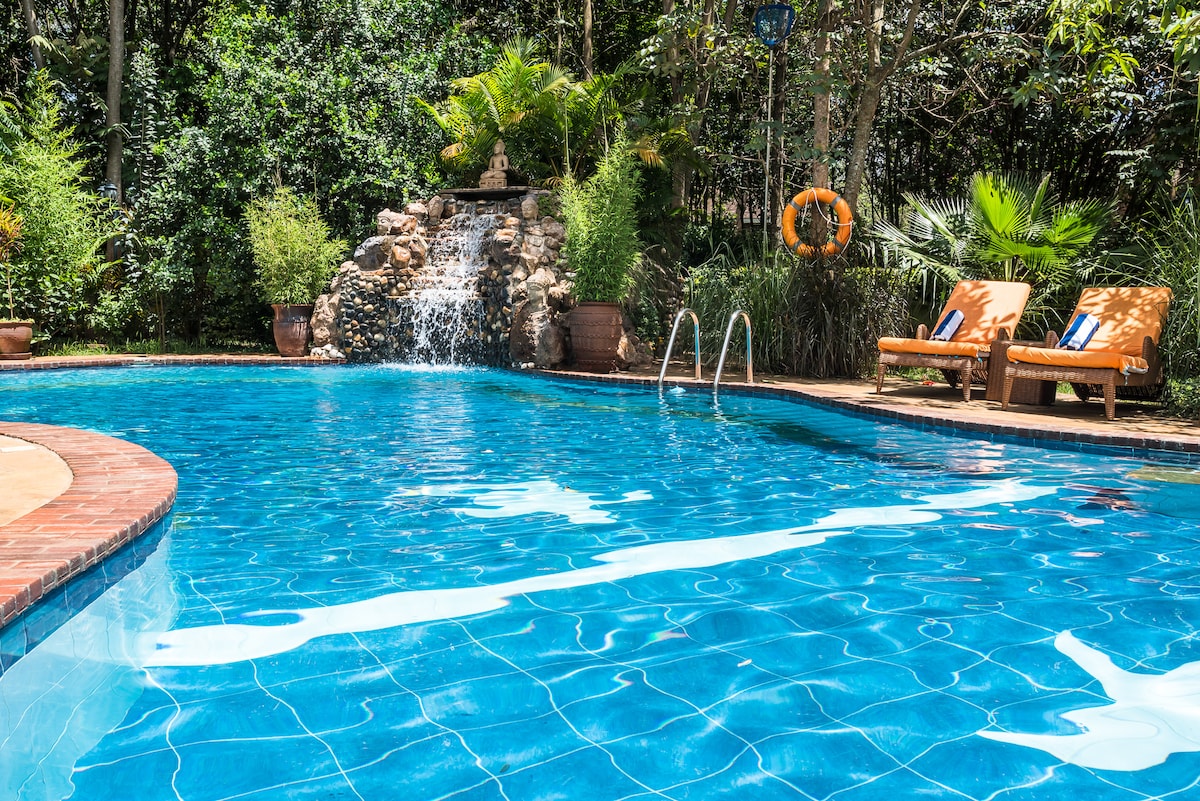 A large swimming pool is surrounded by lush greenery, featuring a waterfall at one end. Two orange sun loungers with blue cushions are positioned nearby, offering a relaxing space for guests to unwind.