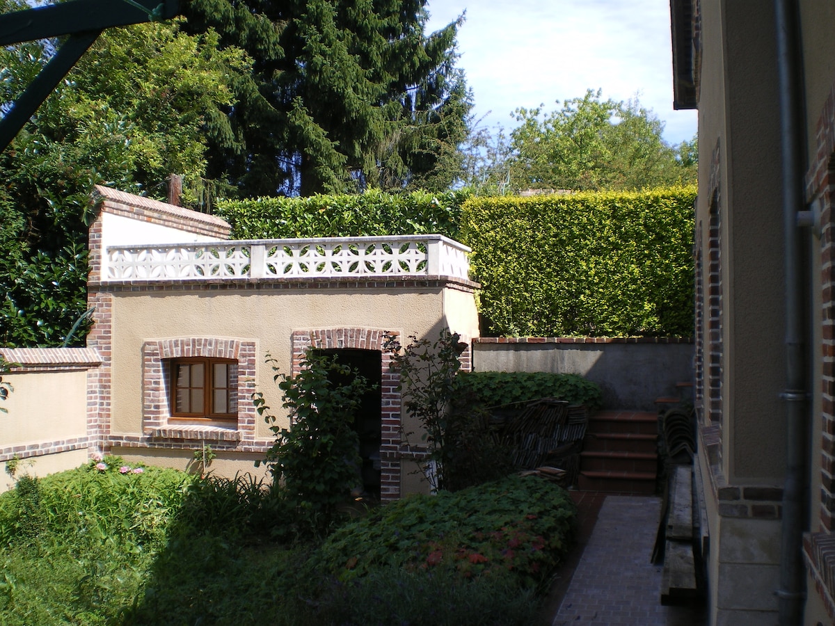 A lush outdoor area is surrounded by greenery, featuring a well-maintained garden with colorful flowers. A partially visible building with a decorative railing sits in the background, while a set of stairs leads to the entrance of the property. The space is bathed in natural light.