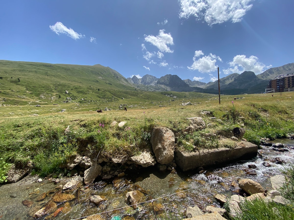 A serene landscape displays rolling green hills under a clear blue sky. Mountains are visible in the background, creating a stunning backdrop. A small stream flows through the foreground, accompanied by scattered rocks and patches of wildflowers, inviting a sense of calm in the natural setting.