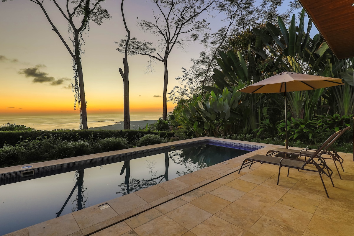 A private pool is highlighted against a backdrop of lush tropical greenery and a colorful sunset over the ocean. Two lounge chairs sit beside the pool, with an umbrella providing shade. The serene environment invites relaxation and connection with nature.