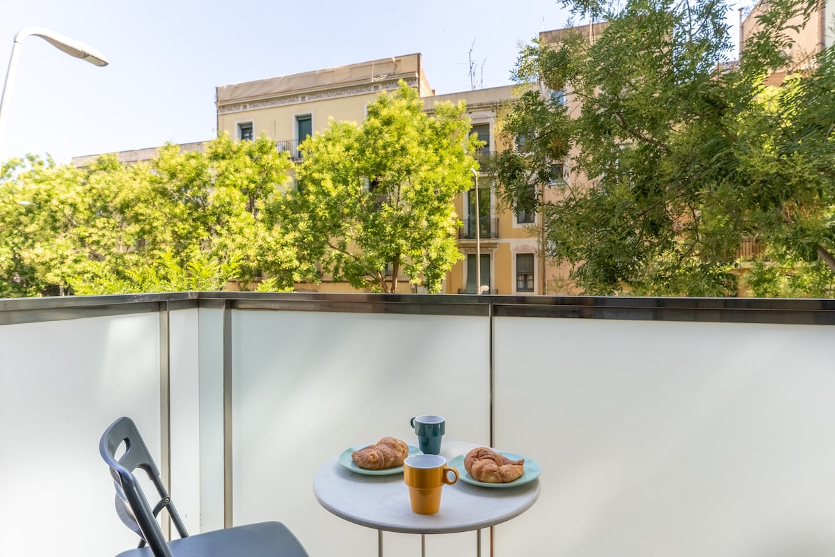 A balcony is seen with a small round table set for two, featuring two croissants and three colorful cups. Lush green trees frame the view, and the building facade is partially visible in the background, creating a serene outdoor setting.