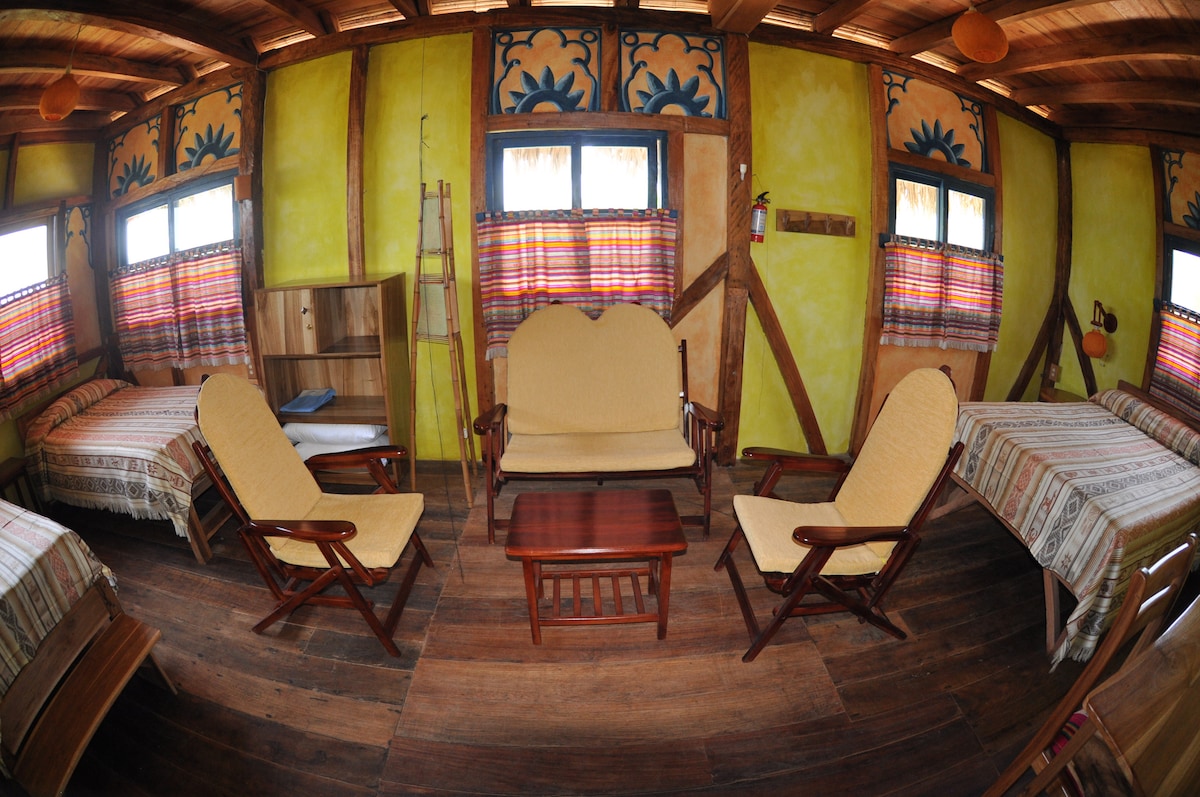 A cozy interior features warm yellow walls and wooden accents. Two beds are visible, along with a comfortable seating area that includes a wooden coffee table and chairs. Large windows adorned with colorful fabric provide natural light to the space.