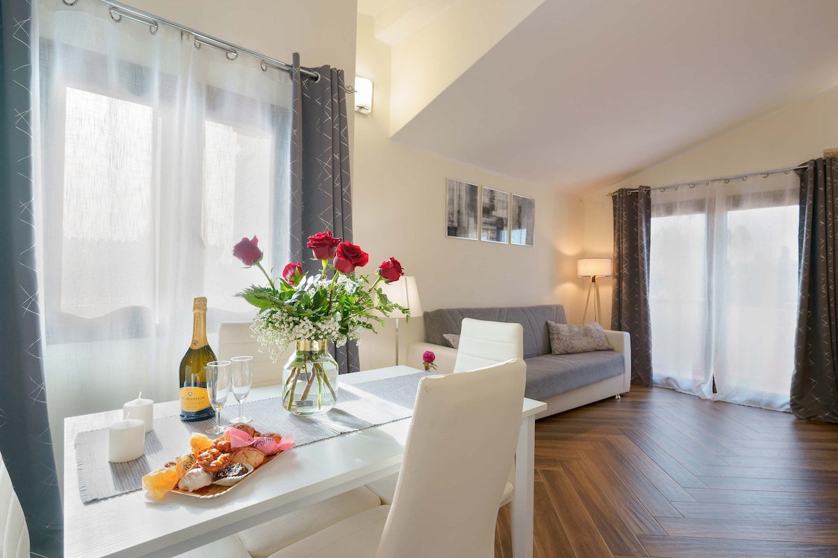 A bright living area features a light gray sofa, a dining table set for two, and elegant floral arrangements. Large windows with soft drapes allow natural light to brighten the space, while a bottle of sparkling wine and glasses sit atop the table, creating an inviting atmosphere.