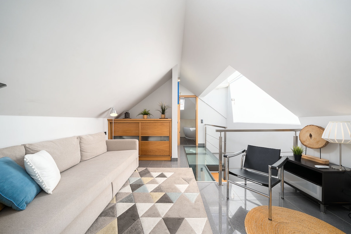 A bright loft area features a spacious, neutral-toned sectional sofa with a blue accent pillow. A geometric patterned rug covers the floor, complemented by a sleek black armchair and a modern side table. Natural light filters through a skylight, enhancing the airy feel of the space.