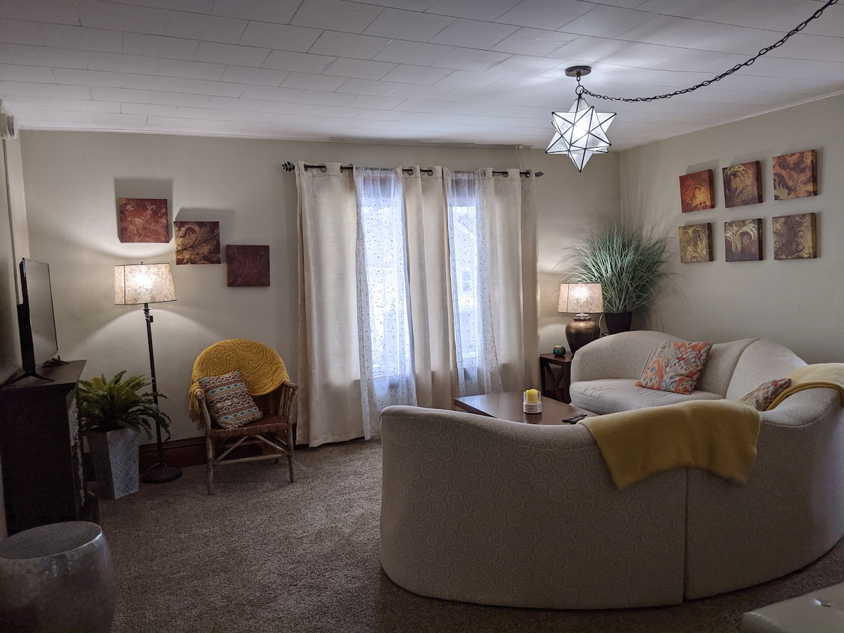 A spacious living room is depicted, featuring a curved sectional sofa and a small side table. Natural light pours in through large curtains, enhancing the soft color palette. A decorative star-shaped light fixture hangs from the ceiling, complemented by two lamps beside the seating area.