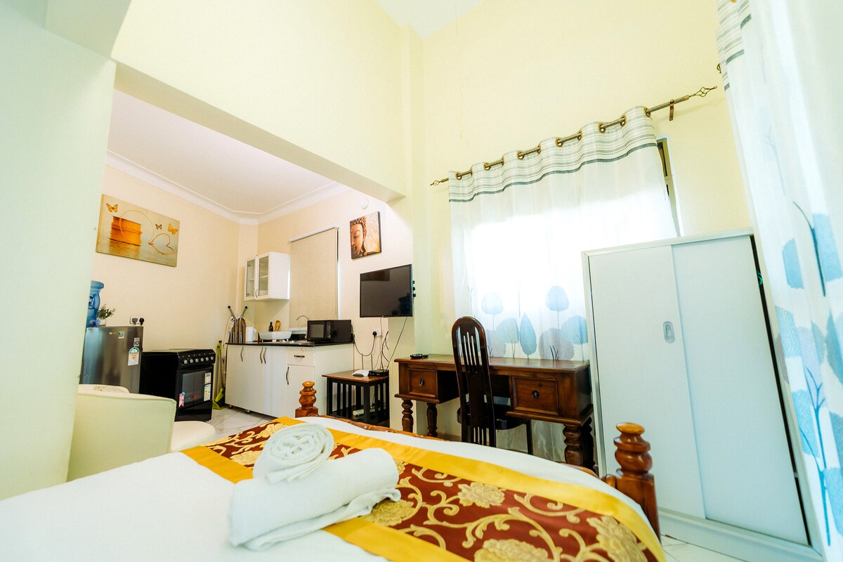 A bright studio space features a compact kitchen, a wooden desk, and a cozy sleeping area. Natural light enters through window treatments, highlighting the white cabinetry. A neatly arranged towel is placed on the spacious bed, complementing warm color accents throughout the room.