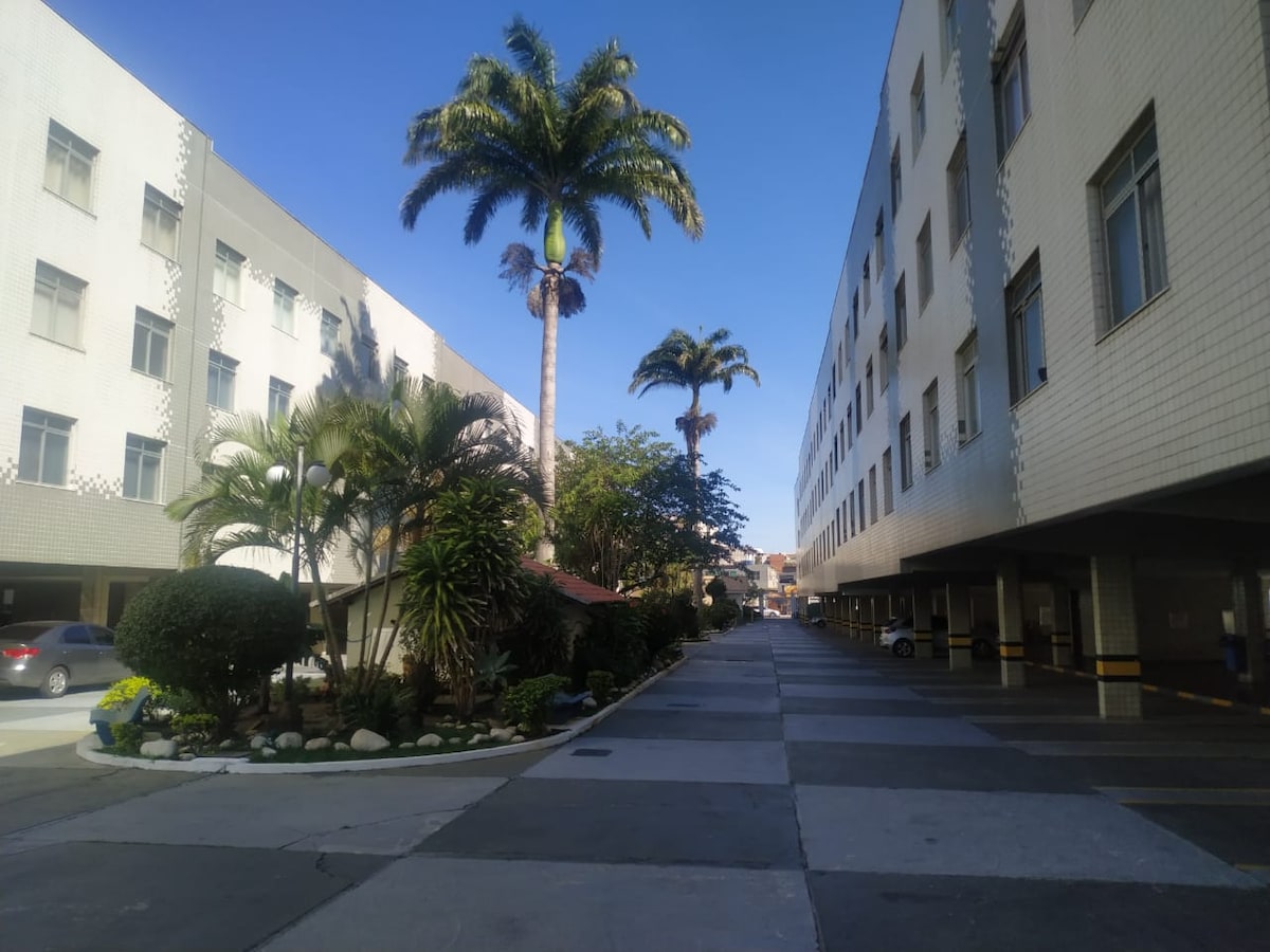 A wide view of a condominium's exterior pathway featuring palm and leafy trees. The neatly paved path leads to the entrance of the buildings, surrounded by bushes and landscaping. The building facades display a clean design, with parking spaces visible to the side.