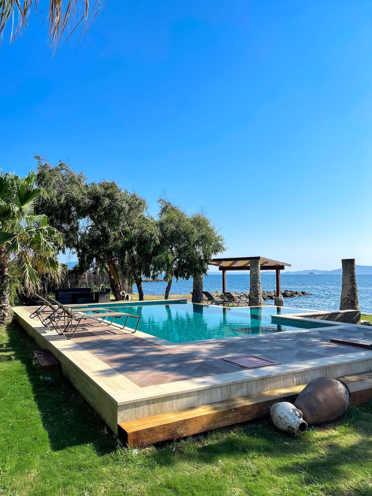 A private swimming pool glistens in the sunlight, surrounded by well-maintained grass and palm trees. Lounge chairs are arranged beside the pool area, and a wooden pergola stands nearby, providing shaded seating with scenic views of the sea.