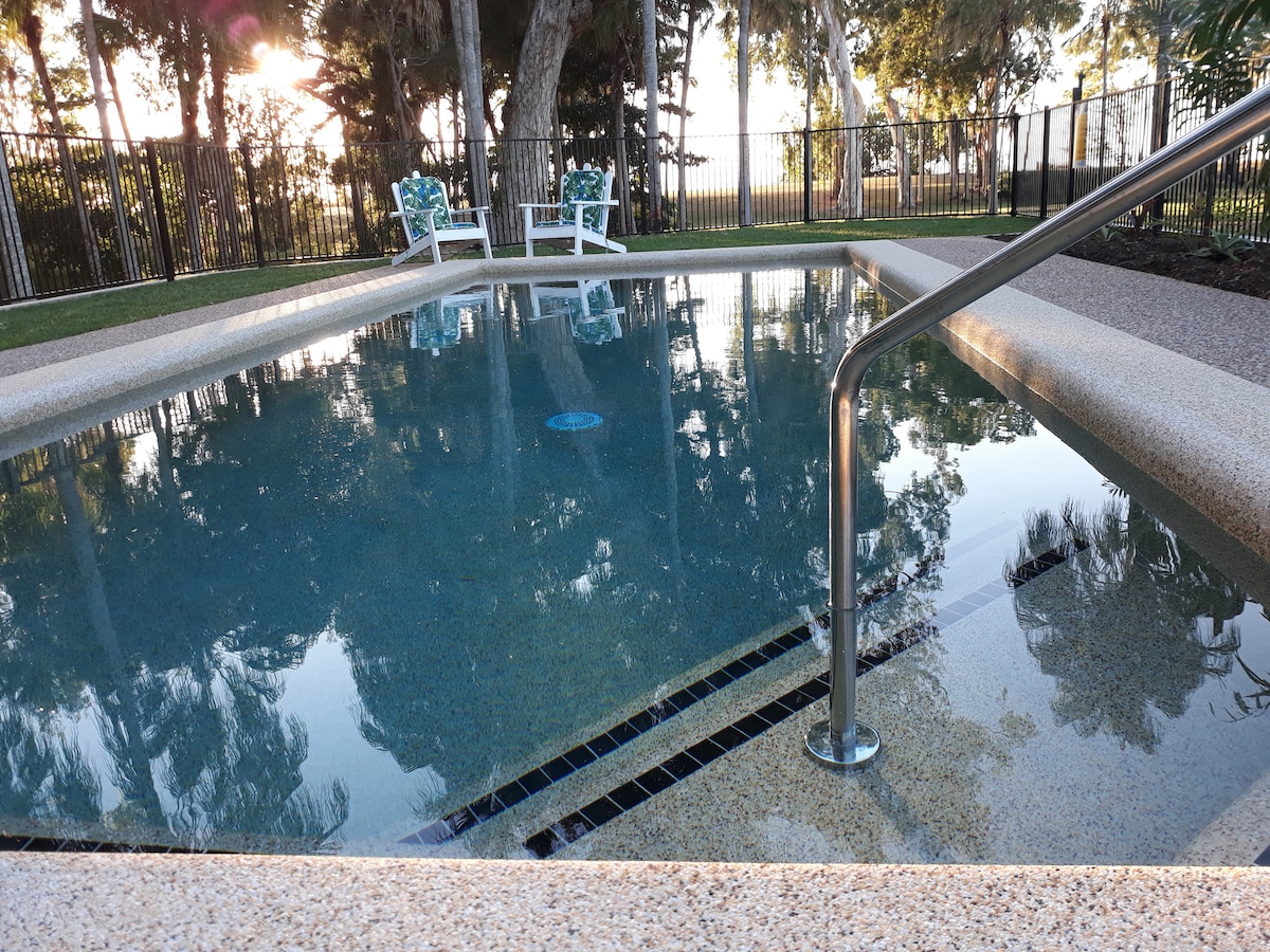 A magnesium pool with a clear, reflective surface is surrounded by neatly manicured grass and a secure fence. Two white outdoor chairs rest beside the pool, offering a serene spot for relaxation. Sunlight filters through nearby trees, casting gentle shadows on the pool area.