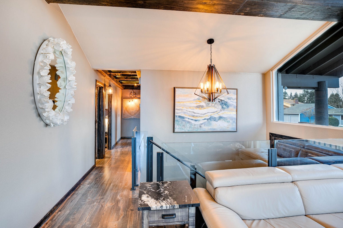 A modern common area is highlighted by a light fixture above and a decorative mirror on the wall. Neutral wall colors complement a large artwork depicting subtle ocean waves. A beige sectional sofa is positioned near a glass railing, providing an open and airy feel.