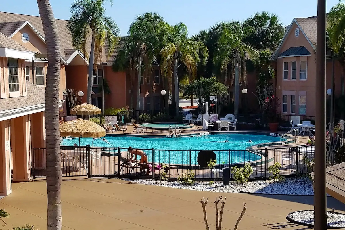 An inviting pool area is surrounded by lush palm trees and tropical landscaping. Lounge chairs and shaded umbrellas offer comfortable seating options. The pool is centrally located within a gated space, with buildings flanking either side, enhancing the community feel of the environment.