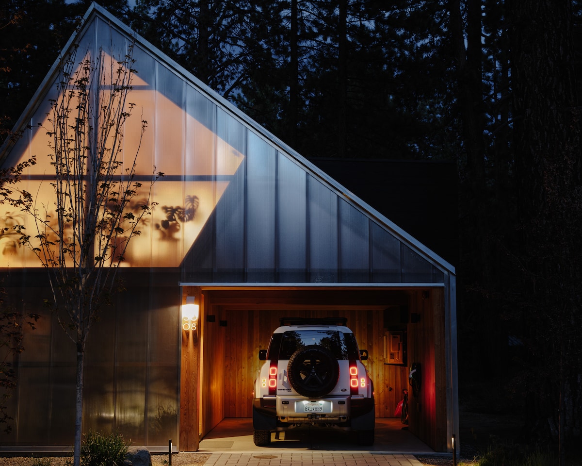 A modern cabin exterior is illuminated at night, featuring a sharp angular roof. A vehicle is parked in the open garage, surrounded by a landscaped area with minimal lighting. Warm light emanates from the glass-walled section, contrasting with the dark forest backdrop.