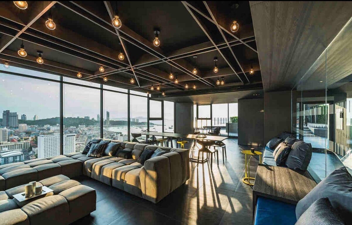 A modern common area features large windows offering panoramic views of the city. Comfortable seating is arranged to encourage relaxation, illuminated by industrial-style pendant lights. Dining tables are present, complemented by a sleek design, creating a welcoming space for social gatherings.