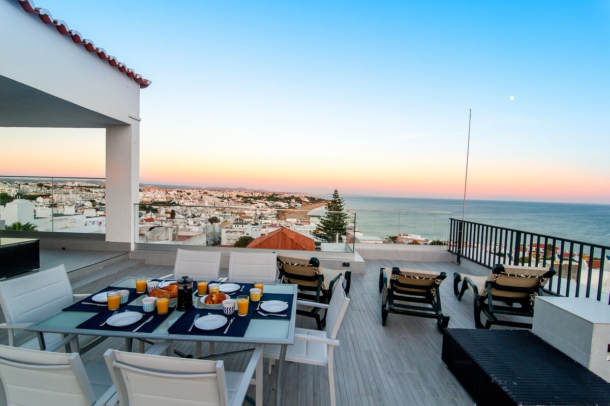 A spacious outdoor terrace presents dining arrangements for six, featuring a glass table set with plates and drinks. Sun loungers are positioned for relaxation, overlooking a panoramic view of Albufeira's skyline and the sea at sunset, with soft hues of orange and blue lighting the sky.