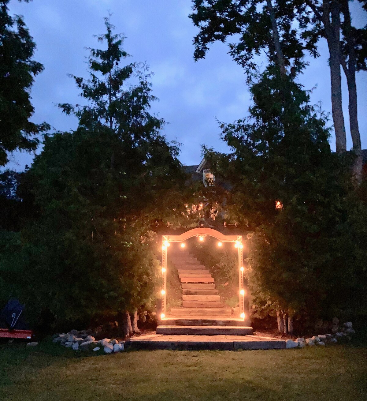 The entrance to the loft is framed by tall, lush trees adorned with soft, glowing lights. A stone pathway leads up the stairs to the entrance, creating a welcoming atmosphere during twilight.