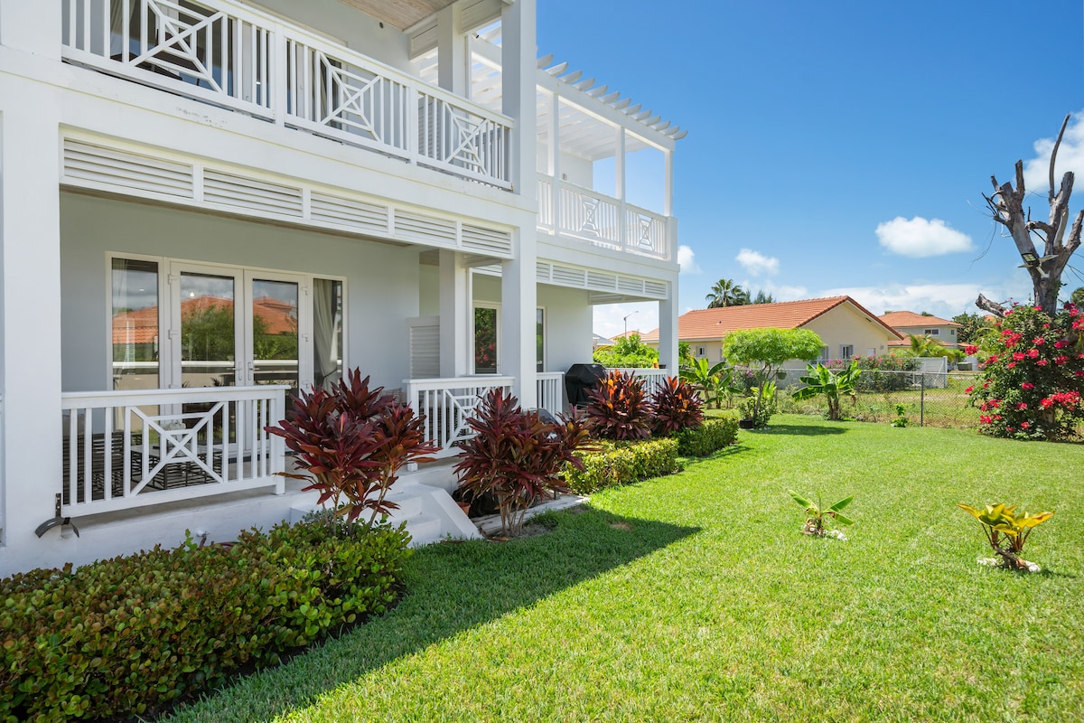 A spacious exterior view showcases a well-maintained garden with lush green lawns and colorful shrubs. A balcony extends from the house, providing an area for outdoor relaxation. The sky is clear and blue, enhancing the serene atmosphere of the residence.