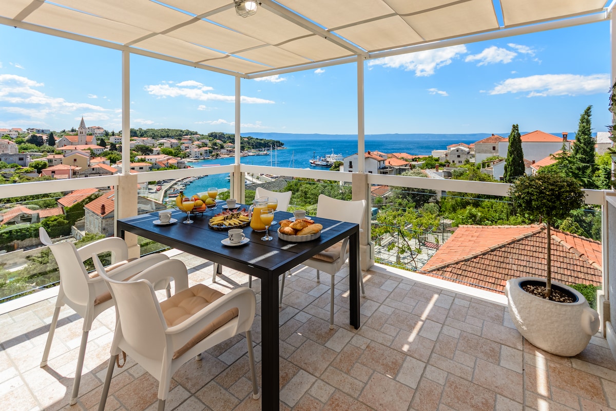 A spacious terrace is set with a dining table, adorned with a variety of breakfast items and drinks. Scenic views of the sea and surrounding rooftops are visible in the background, framed by a bright blue sky and scattered clouds.
