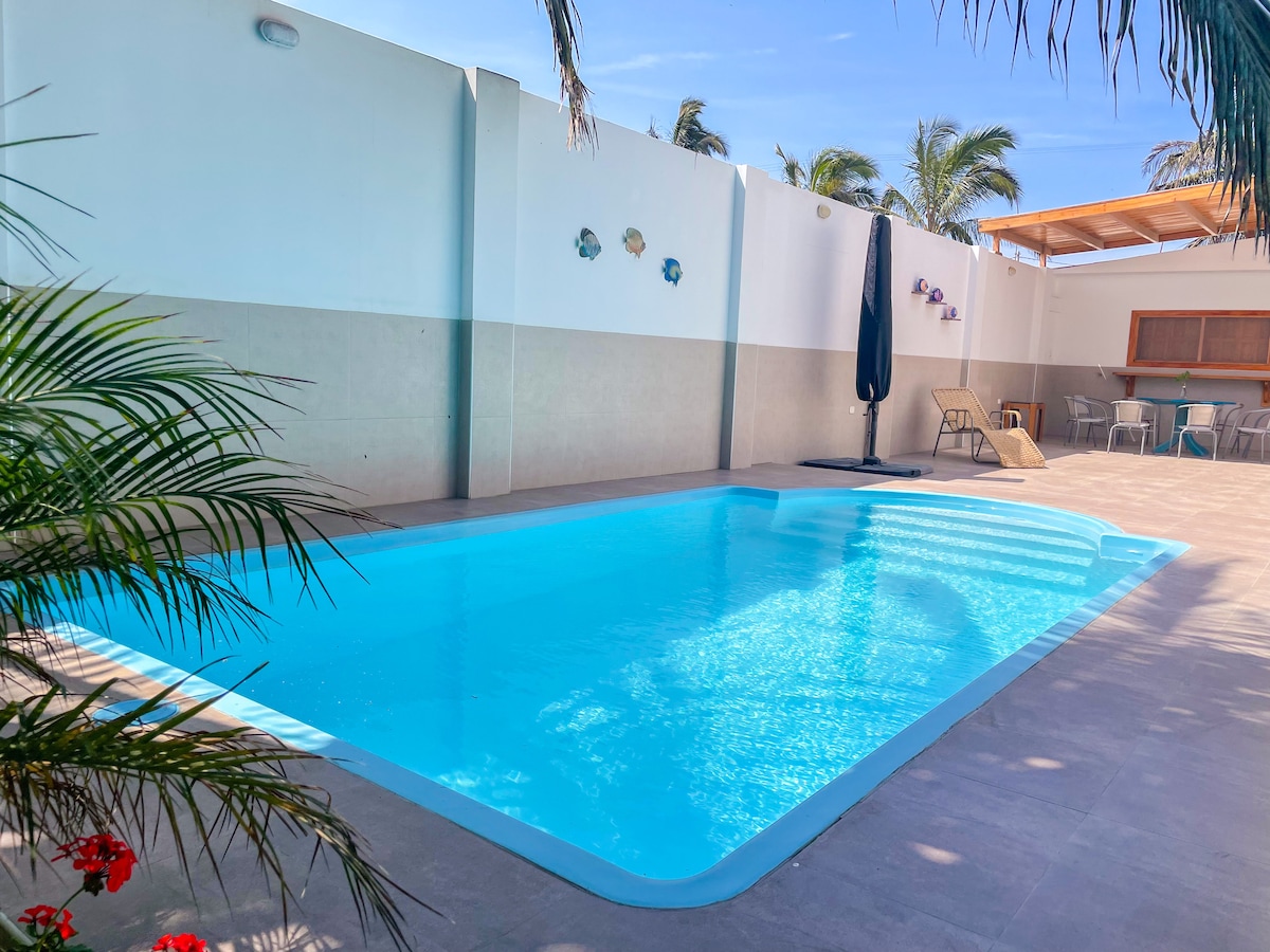 A bright pool area showcases a clear blue swimming pool surrounded by palm trees and tiled flooring. Beach chairs are positioned nearby, and a shaded seating area offers additional relaxation space. The walls are adorned with decorative elements, contributing to a cheerful atmosphere.