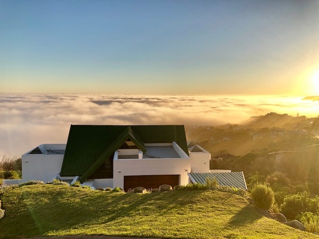 The house features a green roof and white exterior, situated on a hillside above a sea of clouds. A glowing sunset casts a warm light over the landscape, highlighting the serene natural setting and expansive views over the horizon.