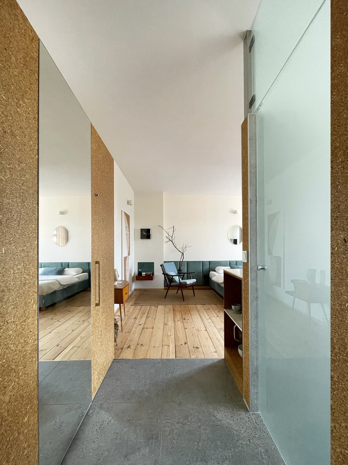 A modern entryway is displayed, featuring a large mirror and a cork panel beside a glass shower enclosure. The open space leads to a bedroom with muted tones, showcasing a comfortable bed and a vintage chair, complemented by light wooden flooring.