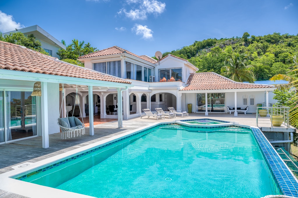 The exterior of a spacious villa is showcased, featuring a large swimming pool with clear blue water. Comfortable lounge chairs are arranged beside the pool. The villa is surrounded by lush greenery and offers covered terraces for relaxation. A well-maintained roof and warm-toned stonework reflect the sunny ambiance.