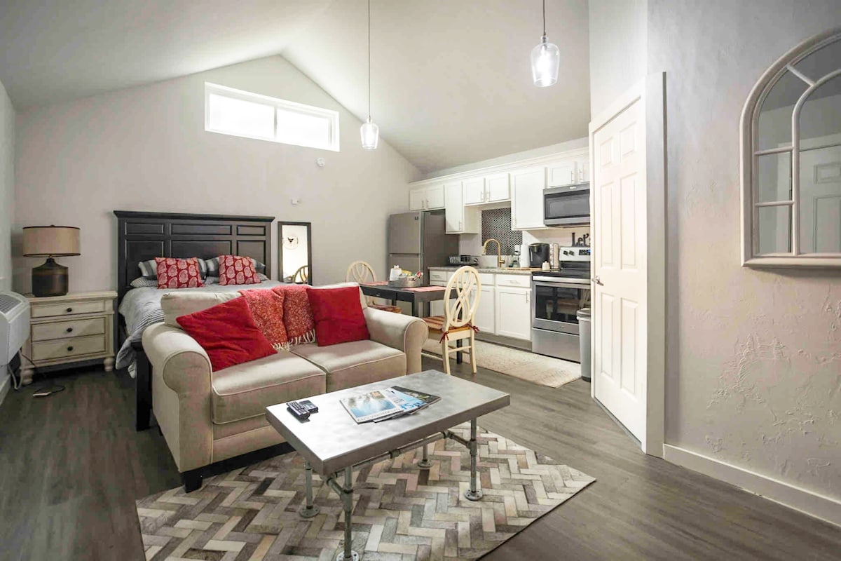 A studio space is revealed, featuring a queen-size bed with decorative pillows and a living area with a comfortable sofa. A dining table with chairs is positioned nearby, and the kitchen is equipped with stainless steel appliances, countertops, and an inviting atmosphere enhanced by natural light.