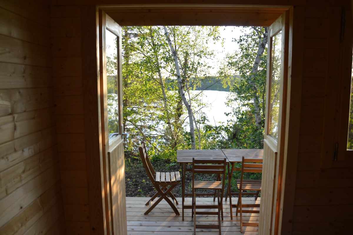 An inviting porch area is visible through double doors, featuring a wooden table and chairs set against a backdrop of lush greenery and the serene lake beyond. Soft natural light filters through the open space, creating a peaceful outdoor atmosphere.