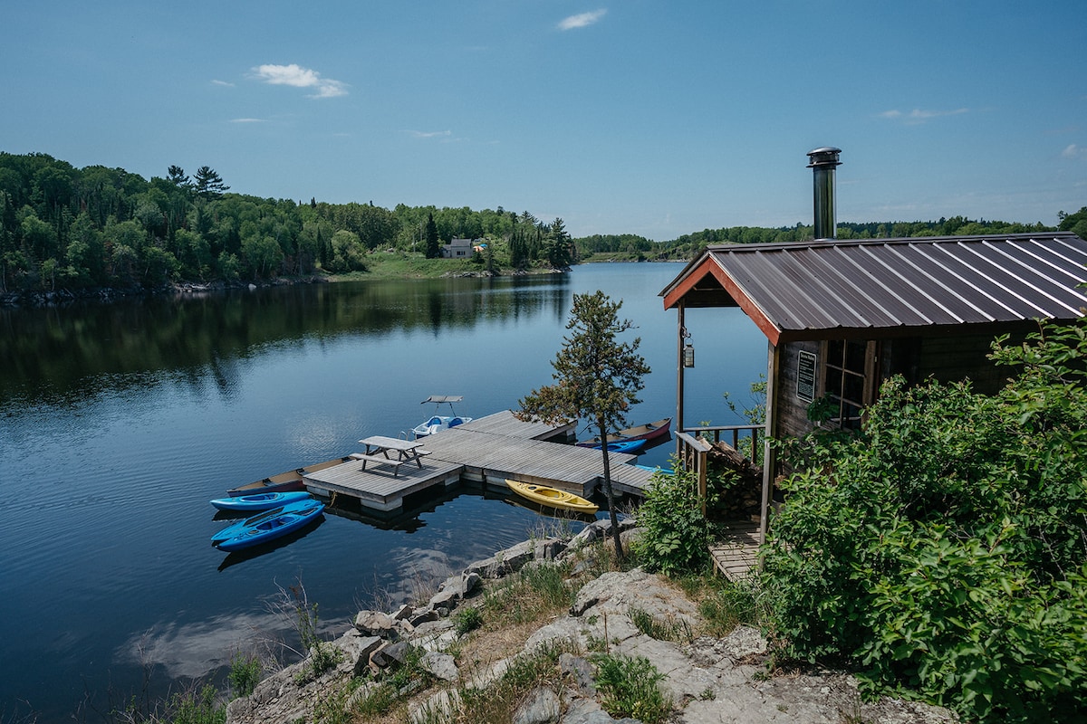 A serene lake view is captured, featuring a wooden dock with canoes and kayaks in vibrant colors. The calm water reflects the greenery and buildings across the lake, while the cabin with a sloped roof sits nestled among rocks, inviting exploration.
