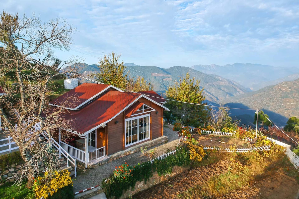 A wooden chalet with a red roof is set against a backdrop of rolling hills. Flowers create a vibrant border around the property, while large windows allow natural light to fill the interiors. The vast landscape stretches into the distance, enhancing the scenic surroundings.