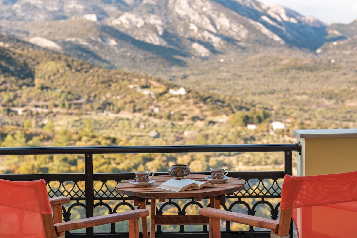 A small wooden table is set for two with orange chairs, overlooking expansive mountain views. Two coffee cups and an open book sit on the table, inviting relaxation. The surrounding greenery and distant hills create a serene backdrop.