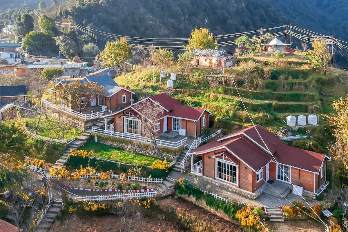 A picturesque view of multiple chalets set against a mountainous backdrop. The chalets feature wooden exteriors with red roofs, surrounded by greenery and pathways. Neatly arranged gardens with colorful plants enhance the outdoor space, while distant hills and power lines form the horizon.