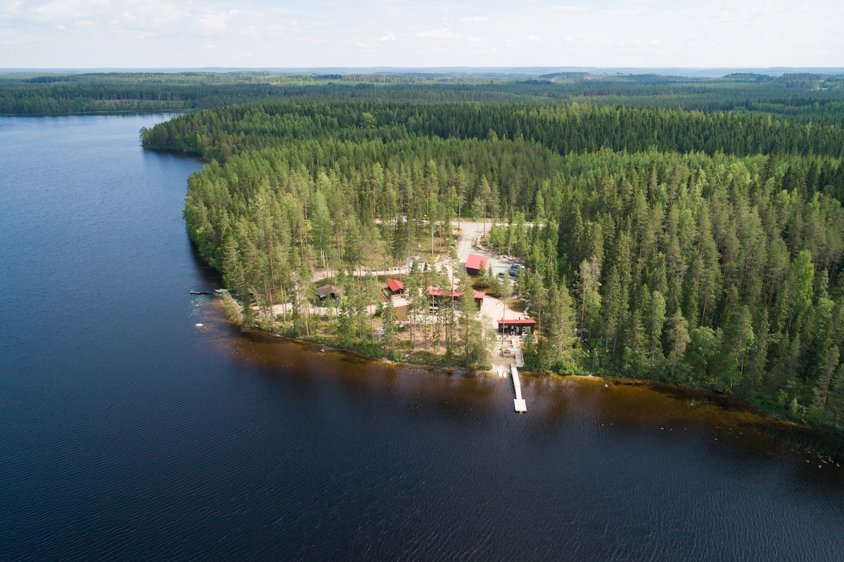 An aerial view showcases the large cabin and surrounding landscape of Central Finland. A private beach is nestled by the calm waters of lake Uuranjärvi, framed by dense greenery, forests, and a wooden dock extending into the lake.