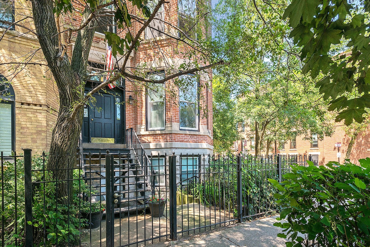 A charming brick building is surrounded by lush greenery and a well-maintained garden. A gated entrance leads to a set of steps that rise to the main entrance. The bright exterior is complemented by large windows, indicating a welcoming atmosphere in a safe neighborhood.