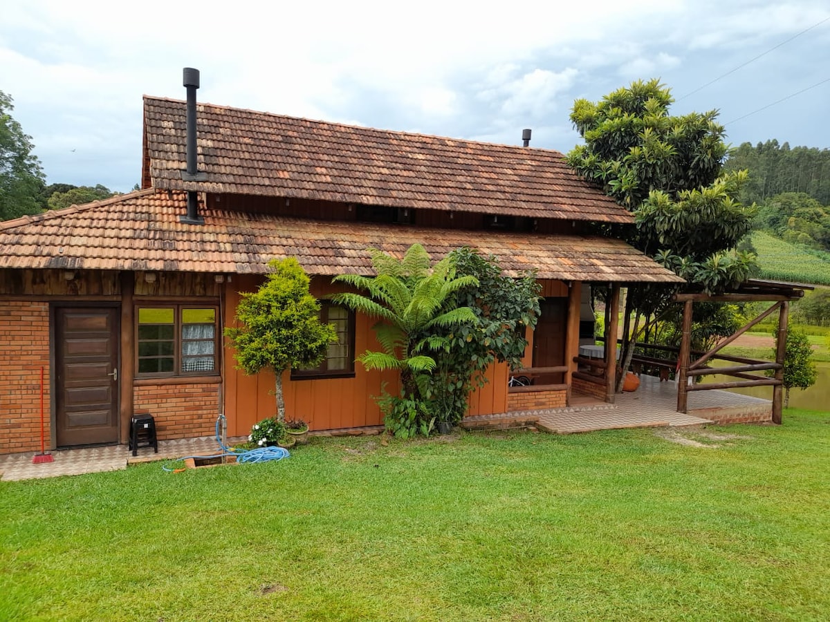 A rustic two-story house features a charming wooden exterior with a sloped roof. The front porch is shaded by trees, while the lush green lawn complements the warm tones of the building. Nearby, a pergola provides an inviting outdoor seating area.