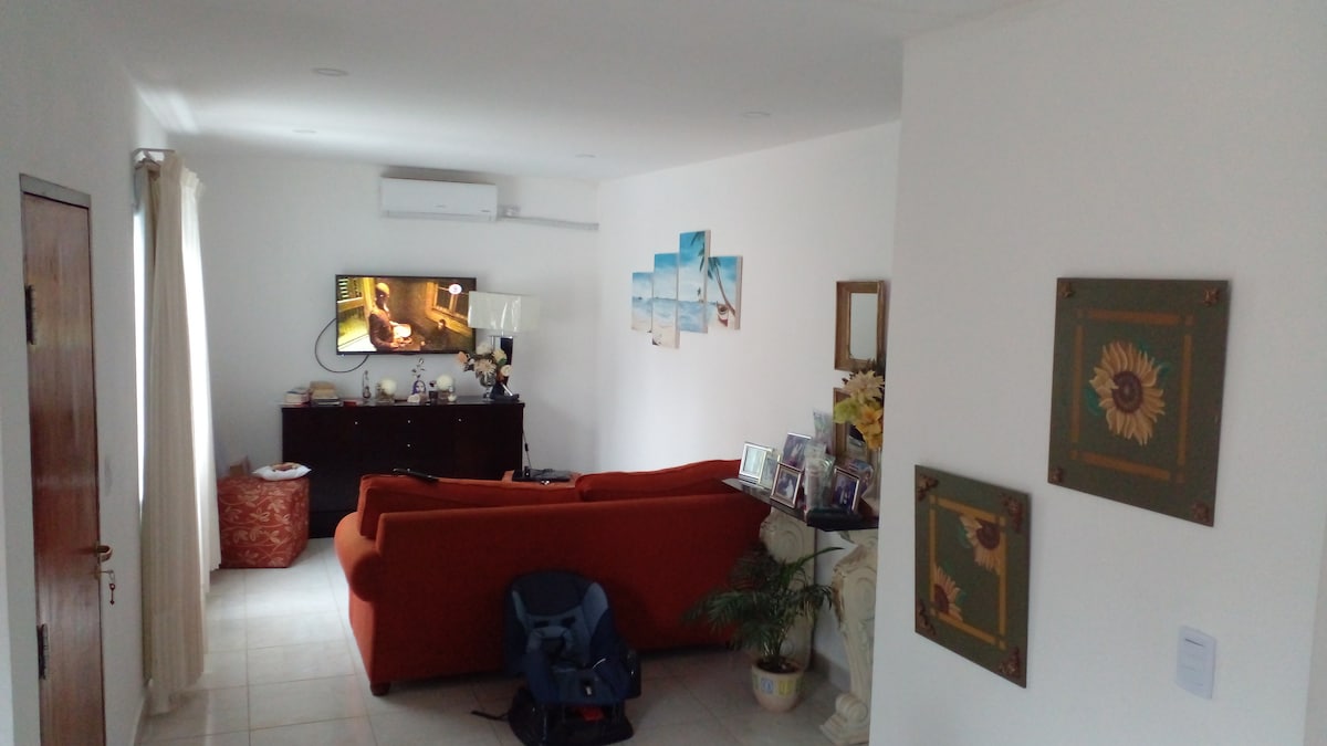 A cozy living area features a bold orange sofa facing a wall-mounted television. Light-colored walls are complemented by decorative paintings. A wooden sideboard holds framed photos, while a potted plant adds a touch of greenery. Sunlight enters through a nearby window, illuminating the space.