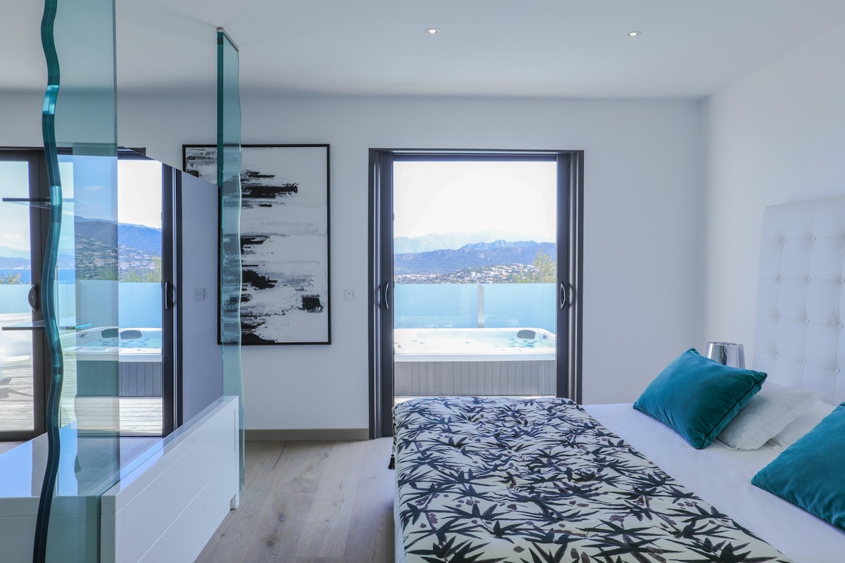 A well-appointed bedroom features a spacious bed with a patterned duvet and decorative cushions. Floor-to-ceiling windows offer unobstructed views of the outdoor terrace and surrounding mountains. A stylish glass partition separates the sleeping area from a private terrace with a jacuzzi.