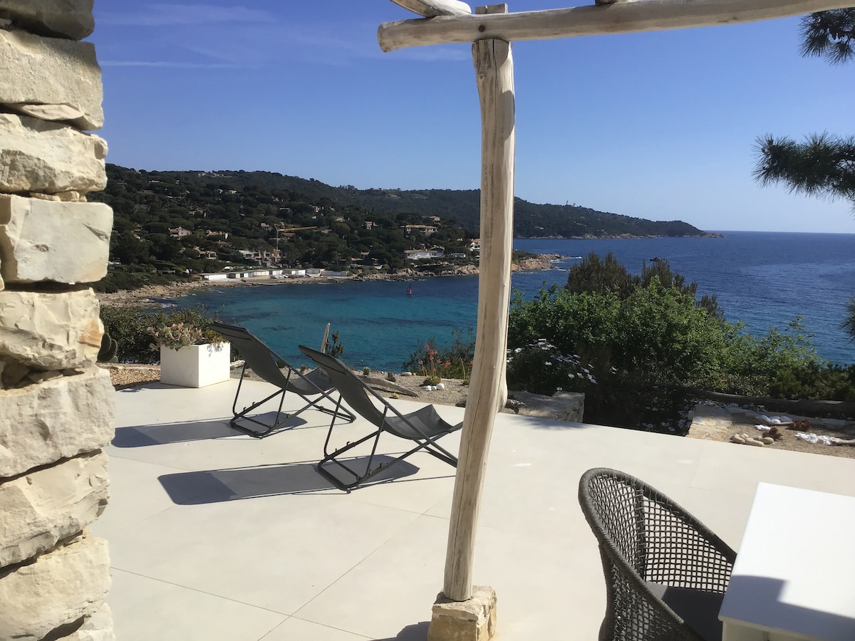 An outdoor terrace is presented, featuring two lounge chairs positioned for optimal sea views. A dining table can be seen in the foreground, complemented by a natural stone pillar that frames the scene. The coastline stretches along the horizon, with varying shades of blue in the water.