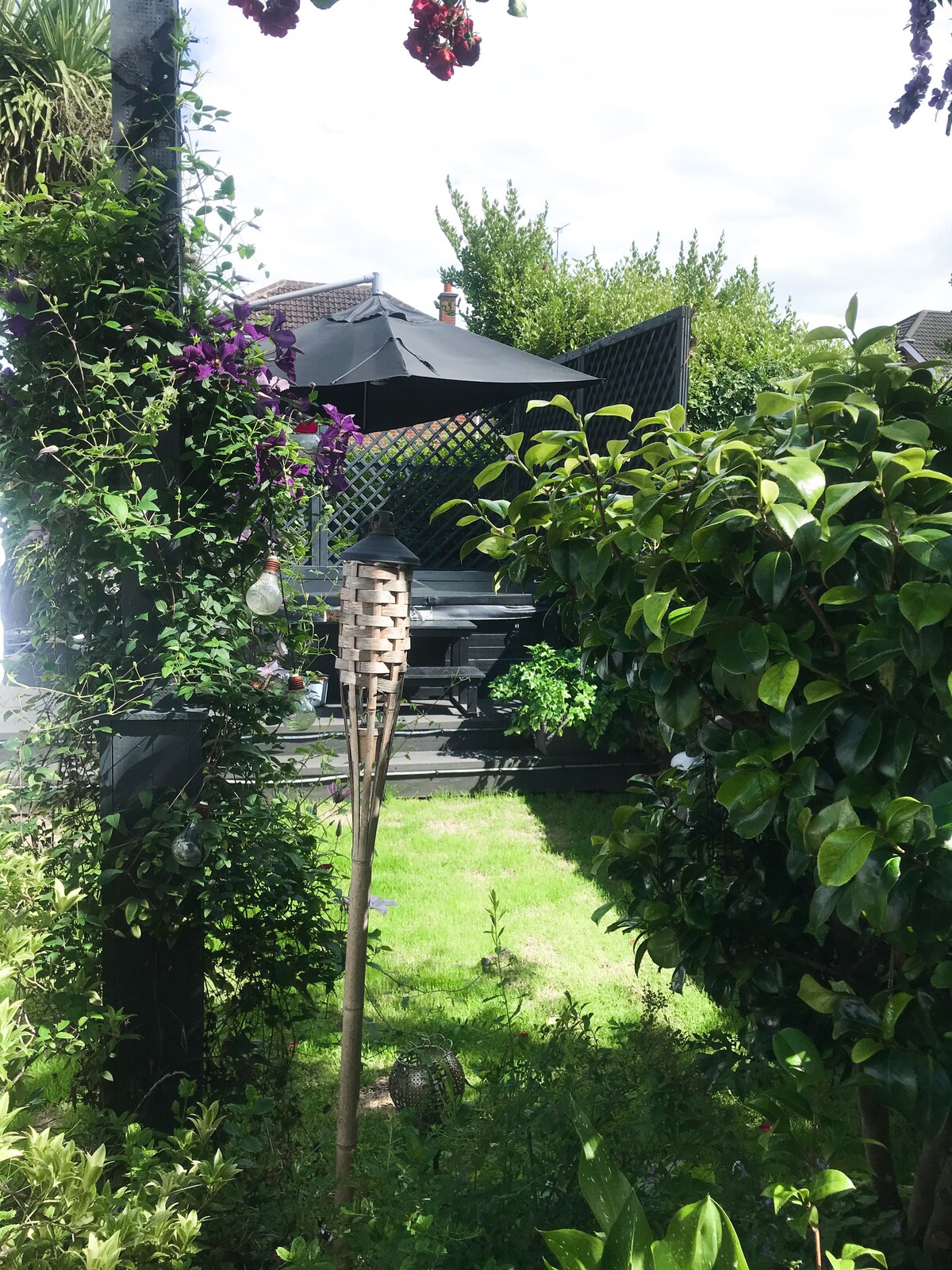 A garden area is surrounded by lush greenery, featuring a mix of flowering plants and shrubs. A patio with a large black umbrella is visible in the background, providing a shaded area for relaxation. Decorative elements, including a bird feeder, enhance the inviting outdoor space.