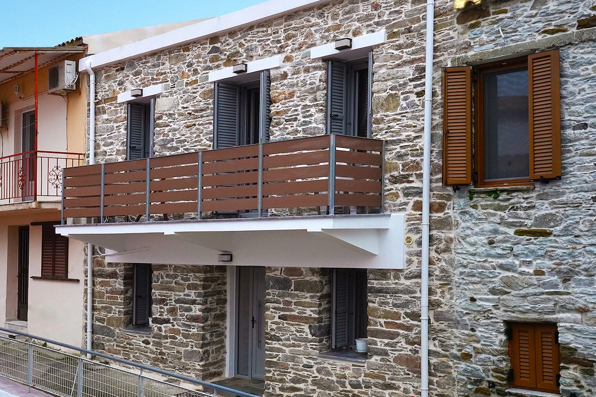 A stone exterior highlights the residence, adorned with wooden shutters and a modern balcony. The balcony features a patterned railing, providing a welcoming outdoor space. The overall facade reflects a blend of traditional and contemporary architectural styles.