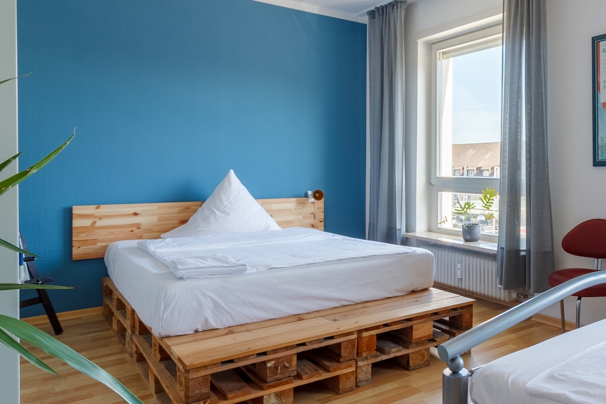 A bright room features a light blue wall and a large window allowing natural light to fill the space. The bed is constructed with wooden pallets, topped with a white duvet. A potted plant and a red chair add vibrant touches to the simplicity of the decor.