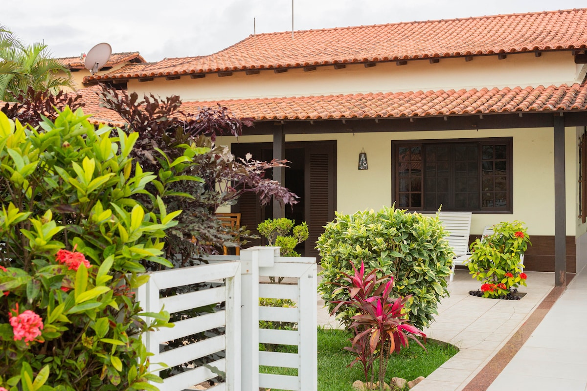 The exterior of the house features a traditional design with a terracotta tiled roof. Lush greenery surrounds the entrance, including vibrant flowering plants and well-maintained hedges, while a white fence provides a welcoming touch.