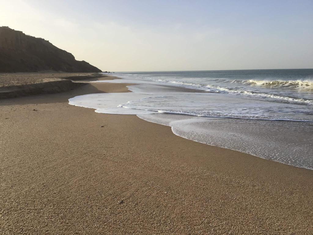 A serene beach stretches along the coastline, with gentle waves lapping at the shore. Soft sand reflects the early morning light, while the horizon meets the calm sea under a clear sky. Rocky cliffs rise in the distance, offering a natural backdrop.
