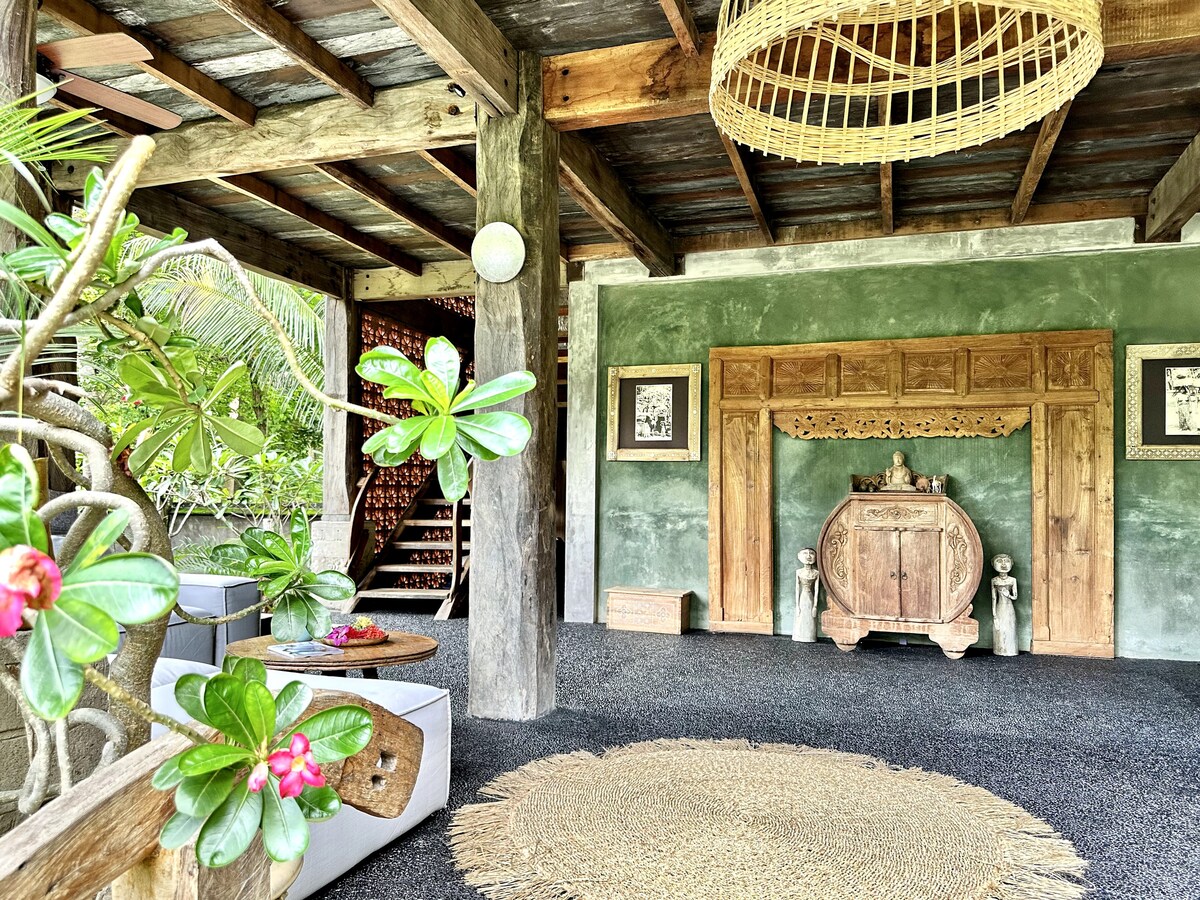 An open living space features a rustic wooden decor, with a large carved wooden cabinet as the focal point. Plush seating is arranged around a natural woven rug, surrounded by greenery. Soft light from a ceiling fixture complements the earthy tones of the walls.