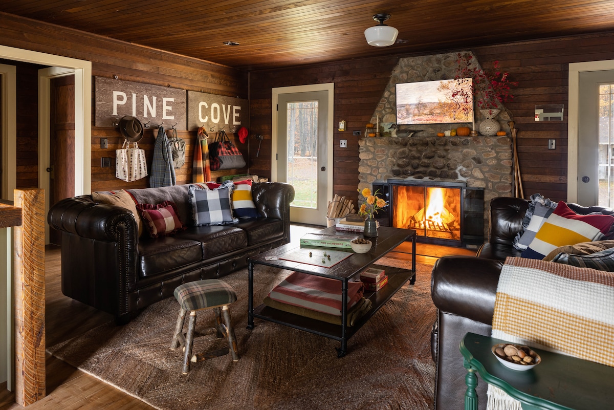 The inviting living area features a stone fireplace with a warm fire, surrounded by dark leather couches and a rustic coffee table. Textiles in earthy tones and a wooden accent wall enhance the cozy ambiance. Natural light enters through nearby doors, creating a welcoming atmosphere.