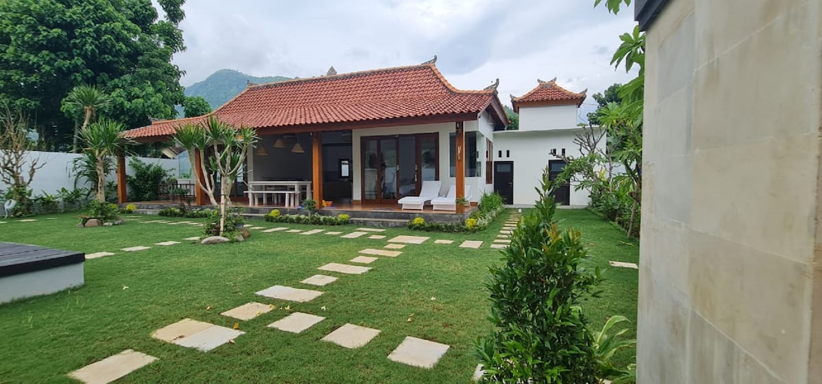 A spacious villa is framed by well-maintained gardens, featuring neatly arranged stepping stones leading to the entrance. The traditional roof design complements the building's exterior. Lush greenery and tropical plants surround the area, enhancing the welcoming environment.
