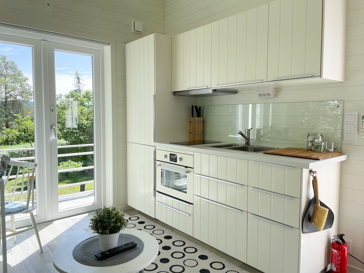 A bright kitchen area features a modern layout with white cabinetry and a countertop. Appliances include an oven, stovetop, and a sink, with natural light streaming through the glass door. A small dining table and chairs are positioned near the door, leading to an outdoor space.